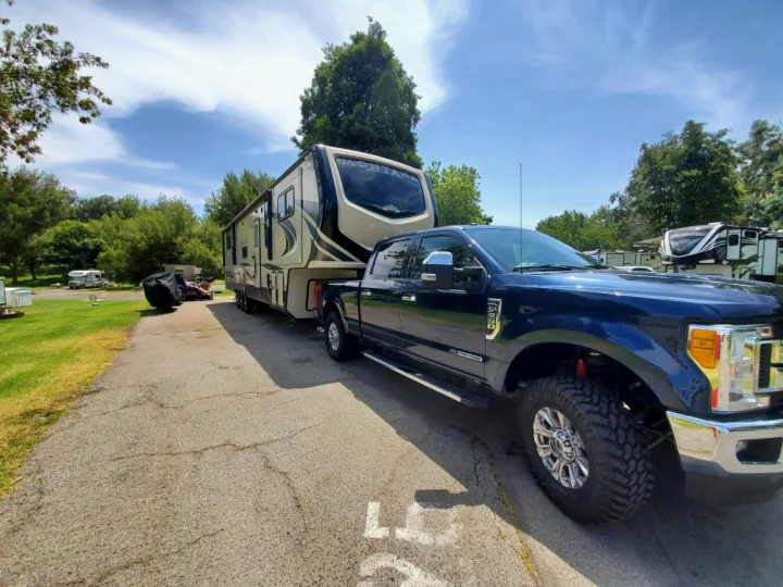 A dark blue pickup truck hitches to a large fifth-wheel RV, ready for an rv rental adventure in a sunny RV park.. Keystone RV Montana High Country Toy Hauler 2019