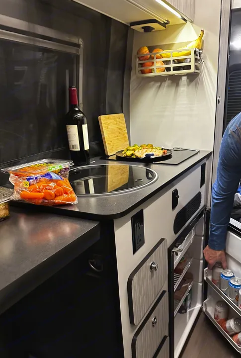 An RV rental kitchen counter with a prepared meal, wine, fruit basket, and a person reaching into a compact fridge.. Winnebago Revel 2020