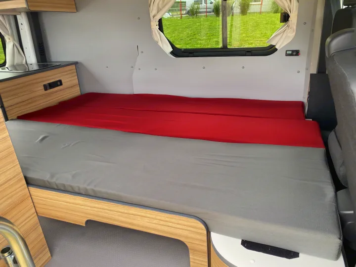 RV rental interior showcasing a cozy, convertible bed with grey and red cushions, wooden cabinetry, and windows, ideal for travel.. Renault TRAFIC 2023