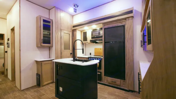 Modern RV rental kitchen featuring a central island with sink, black appliances (fridge, microwave, stove), and light wood cabinetry.. Forest River Cherokee Arctic Wolf 2022