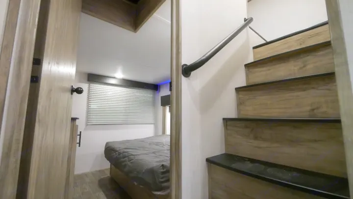 Modern RV rental interior with a cozy bedroom visible through an open door, beside a sleek staircase featuring a black handrail.. Forest River Cherokee Arctic Wolf 2022