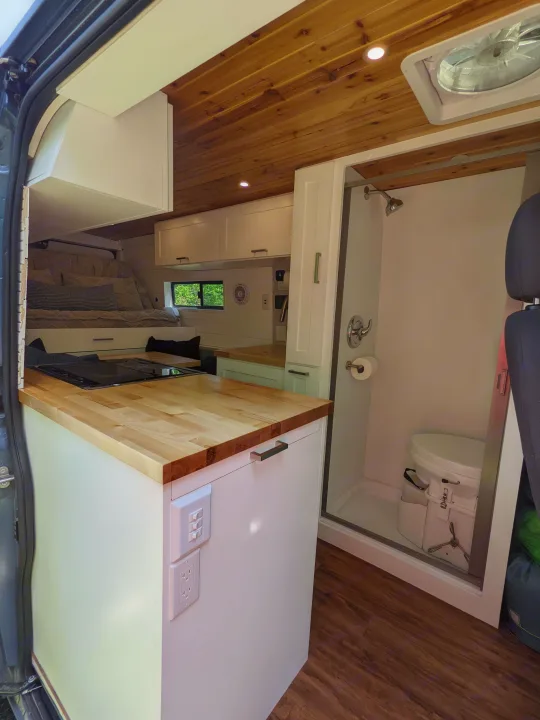 Modern rv rental camper van interior: kitchen with cooktop, cozy bed, wooden ceiling, and a full shower/toilet.. RAM Promaster 2019