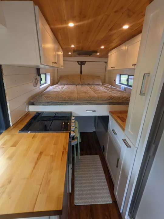Cozy RV rental interior shows a compact bed, integrated kitchen, warm wood ceiling, and white storage cabinets.. RAM Promaster 2019