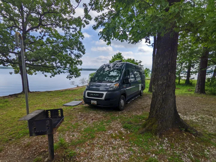 Serene lakeside RV rental spot: a dark camper van under shady trees, next to a grill, by the water.. RAM Promaster 2019