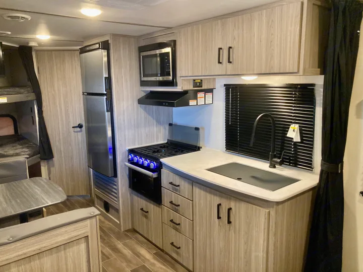 This rv rental interior features a modern kitchen with a stove, sink, fridge, microwave, light wood cabinets, dining area, and bunk beds.. Keystone RV Passport SL 2022