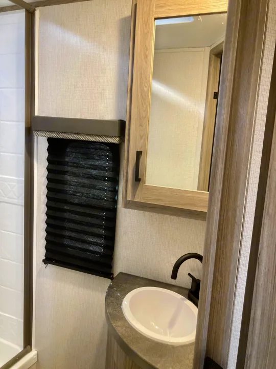 RV rental bathroom with sink, mirror, and shower, featuring modern fixtures and a black pleated window blind.. Keystone RV Passport SL 2022