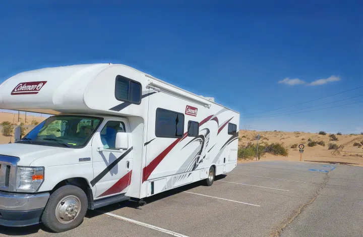 White Coleman RV rental parked in a desert lot, under a blue sky with sand dunes and power lines in the background.. Thor Coleman 2021
