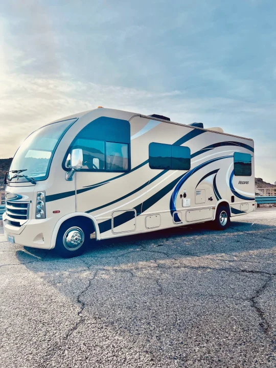 Modern tan Class A Vegas RV rental with black & blue decals, parked on cracked asphalt under a partly cloudy sky.. Thor Vegas 2017