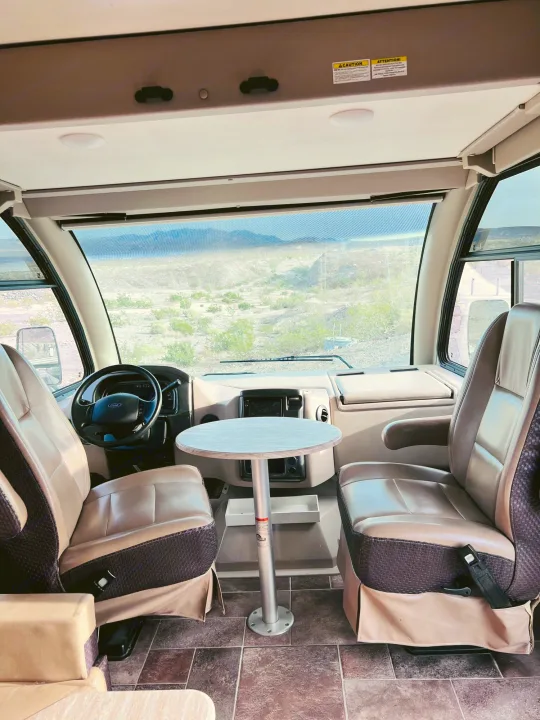 RV rental cabin interior with captain's chairs, table, and a wide desert view through the windshield. Perfect for road trips.. Thor Vegas 2017