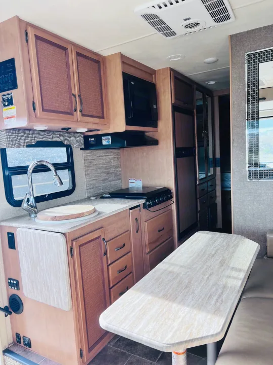 RV rental interior: kitchen with sink, stove, microwave, cabinets, and a light-colored dining table with bench seating.. Thor Vegas 2017