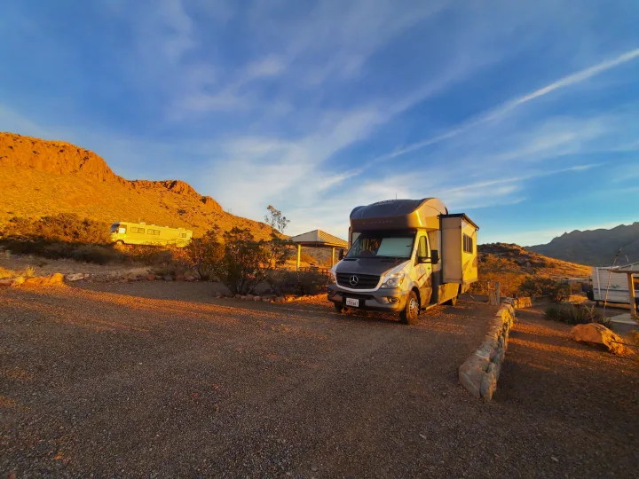 A modern Winnebago RV rental is parked on a gravel site in a desert campground at sunset, with mountains in the background.. Winnebago View 2017