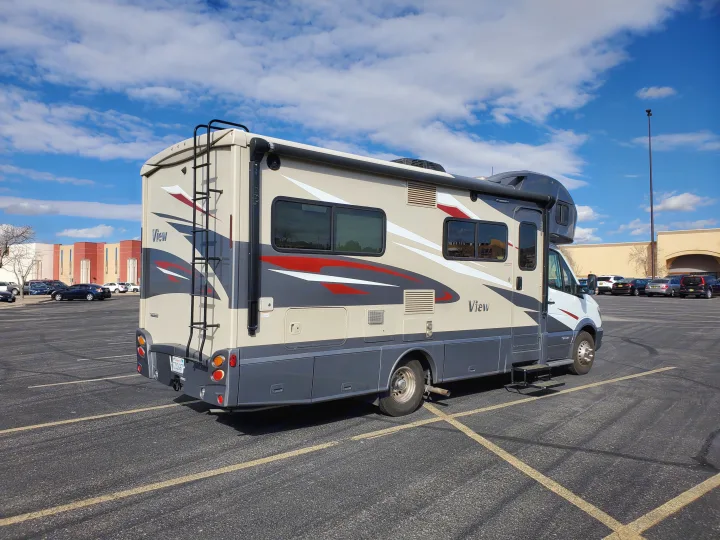 This RV rental, a beige motorhome, is parked in a large asphalt lot. Clear blue sky overhead for travel.. Winnebago View 2017