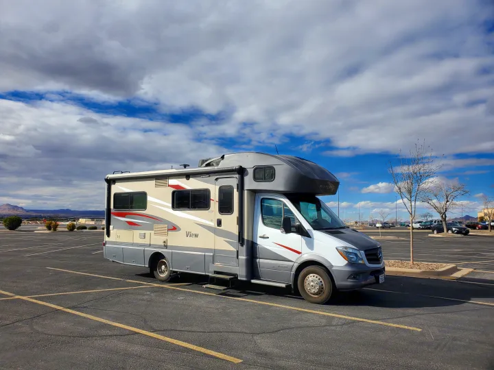 A white and gray Winnebago View RV rental is parked in an asphalt lot under a cloudy sky, ready for travel adventures.. Winnebago View 2017