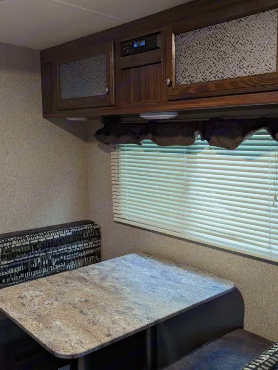 RV rental interior: A cozy dinette with a speckled table, patterned bench seating, and overhead cabinets above a window with blinds.. Coachmen Apex Nano 2019