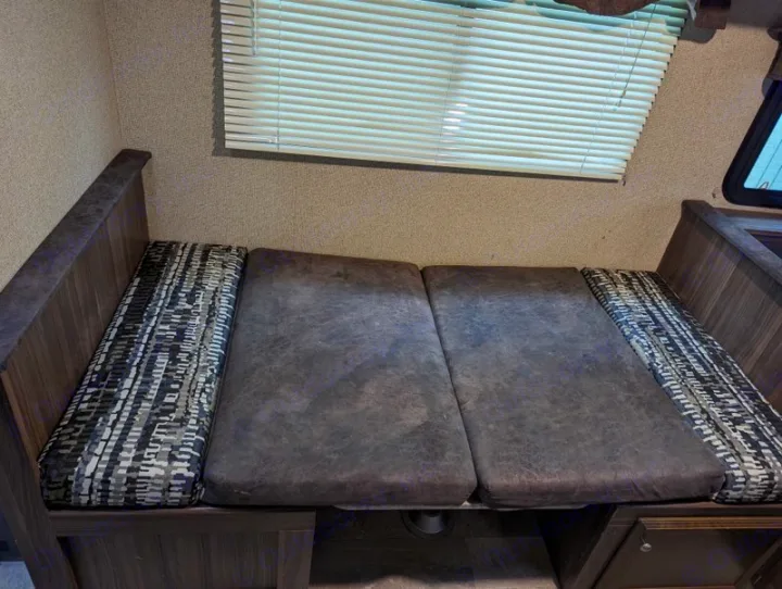RV rental dinette: a cozy seating area with dark grey and patterned cushions, illuminated by a window with blinds.. Coachmen Apex Nano 2019