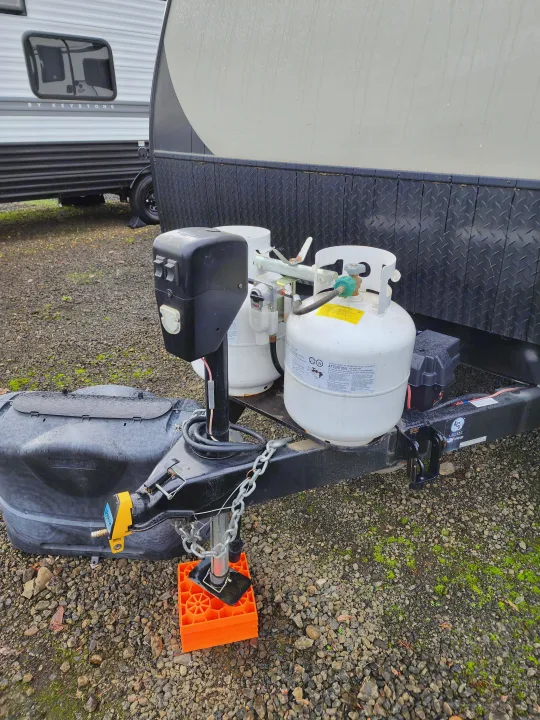 Front of an RV rental travel trailer, showcasing propane tanks, power jack, and hitch setup on a gravel lot.. Keystone RV Passport Grand Touring 2018