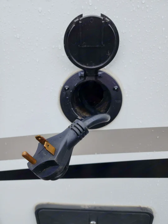 RV rental electrical hookup. A 30-amp shore power plug hangs near the RV's open inlet, ready for connection in the rain.. Keystone RV Passport Grand Touring 2018