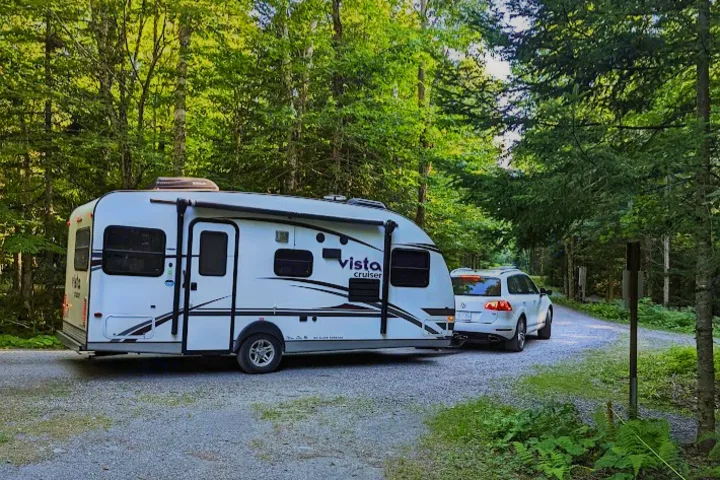 White RV rental travel trailer towed by SUV on a scenic gravel road in a lush forest, perfect for camping.. Gulf Stream Vista Cruiser M-19RSD 2013