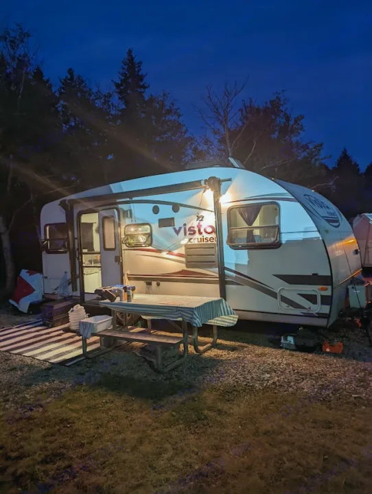 Cozy RV rental at night: A well-lit travel trailer parked at a campsite with a picnic table, surrounded by trees.. Gulf Stream Vista Cruiser M-19RSD 2013