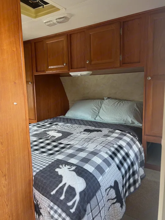 Enjoy comfort in this RV rental bedroom: bed with moose quilt, wooden cabinets, and bright overhead light.. Holiday Rambler Campmaster 2011