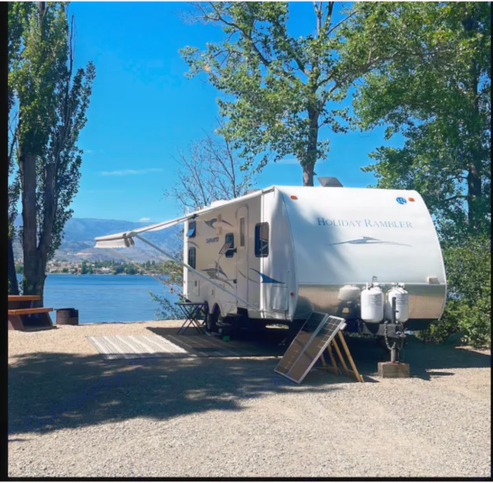 Lakeside serenity: an RV rental (white travel trailer with awning extended) at a gravel site, facing a blue lake & mountains.. Holiday Rambler Campmaster 2011
