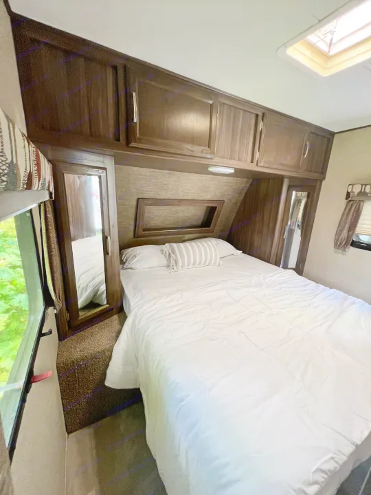 Spacious RV rental bedroom. Features a cozy bed with white linens, wood cabinets, and natural light from a window and skylight.. Coachmen Apex 2018