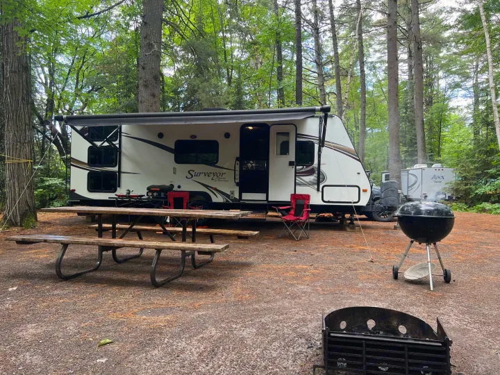 Relaxing RV rental campsite featuring a travel trailer, picnic table, and grill amidst lush trees.. Forest River Surveyor Sport 2014