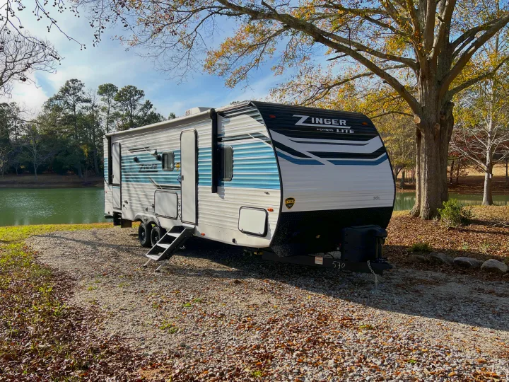 An RV rental, a Zinger travel trailer, parked by a peaceful lake on a sunny autumn day. Perfect for nature escapes.. Crossroads Zinger 2023