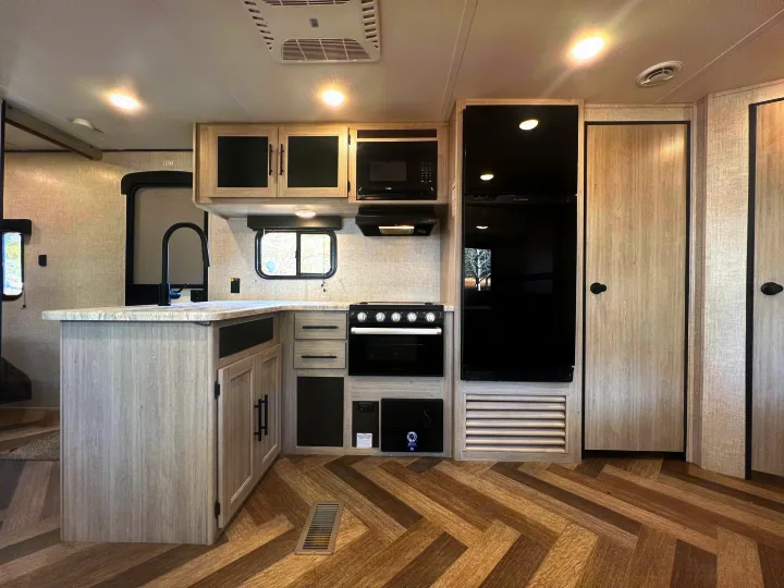 Bright, modern RV rental kitchen featuring light wood-look cabinets, black appliances, and herringbone flooring.. Crossroads Zinger 2023