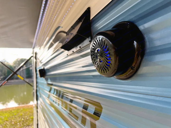 Close-up of an RV rental's exterior, showing a black outdoor speaker with blue LEDs, ribbed siding, & LED awning lights. Parked by a lake.. Crossroads Zinger 2023