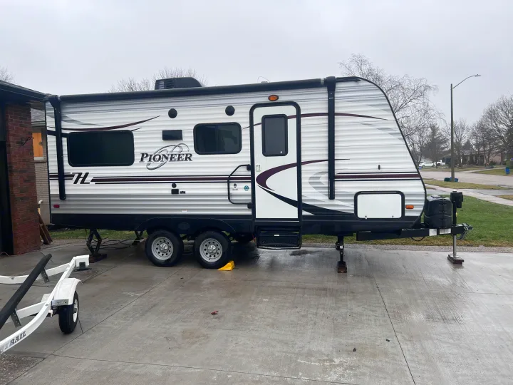 A large white Pioneer travel trailer, parked on a concrete driveway, ready for an rv rental. A boat trailer is nearby.. Heartland RVs Pioneer 2016