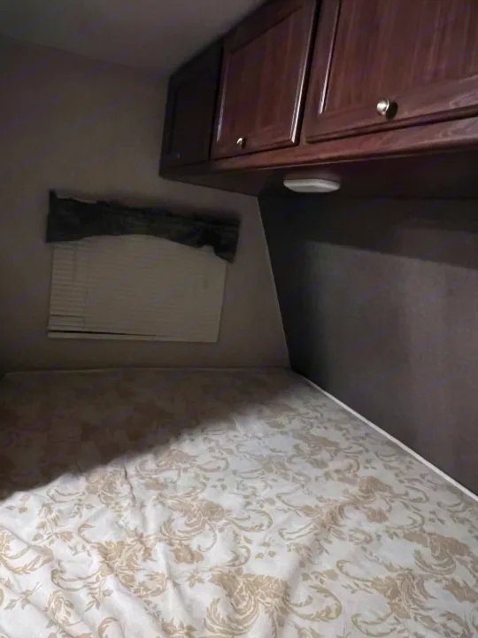 RV rental bedroom: patterned bed, window with blinds, overhead wood cabinets, and light, showing a compact interior.. Heartland RVs Pioneer 2016