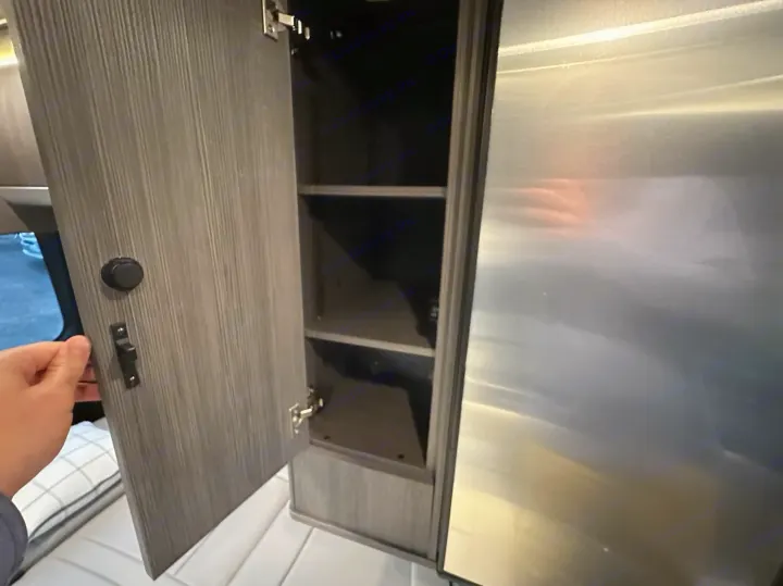 RV rental interior: Hand opens a compact wood-grain cabinet with shelves beside a metal appliance, showcasing smart storage.. Airstream Interstate 2021
