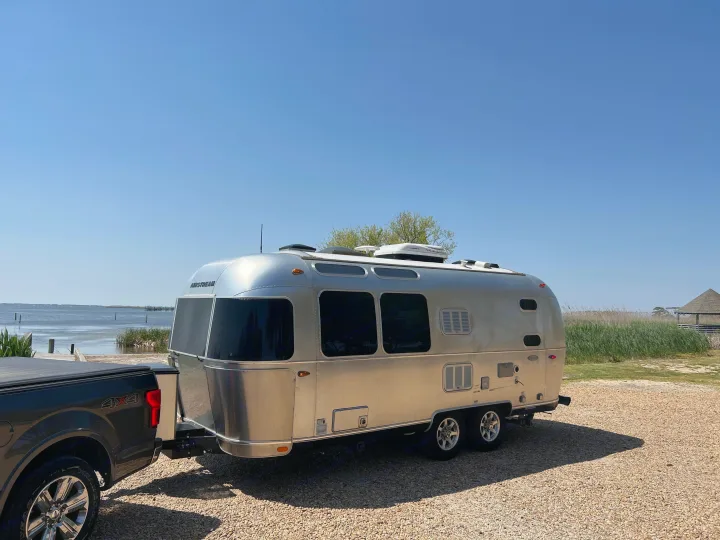 A silver Airstream rv rental, hitched to a pickup truck, parked by a calm bay for serene camping.. Airstream Flying Cloud 2017