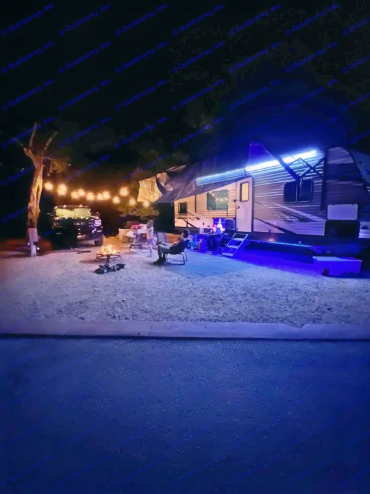 Cozy night at a campground. An RV rental is lit with blue LEDs, complementing warm string lights & a campfire with people gathered.. Heartland RVs Trail Runner 2020