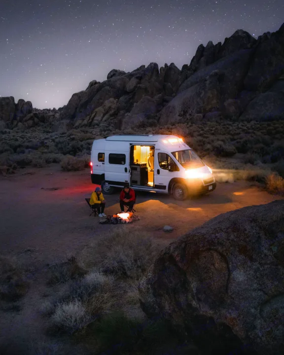 An RV rental adventure under the stars: Two people relax by a glowing campfire, next to their illuminated camper van in a vast, rocky desert.. Winnebago Solis 2021