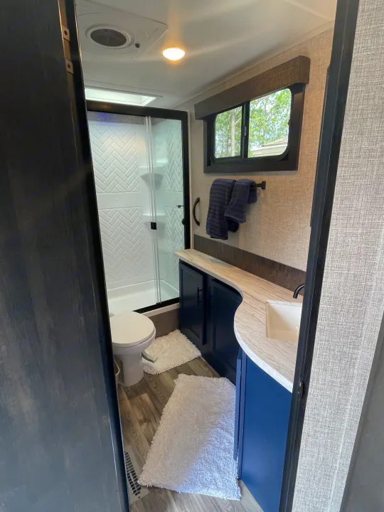 Bright RV rental bathroom with shower, toilet, navy vanity, and window. Includes fluffy mats and dark towels for a cozy travel space.. Jayco Eagle HT 2022