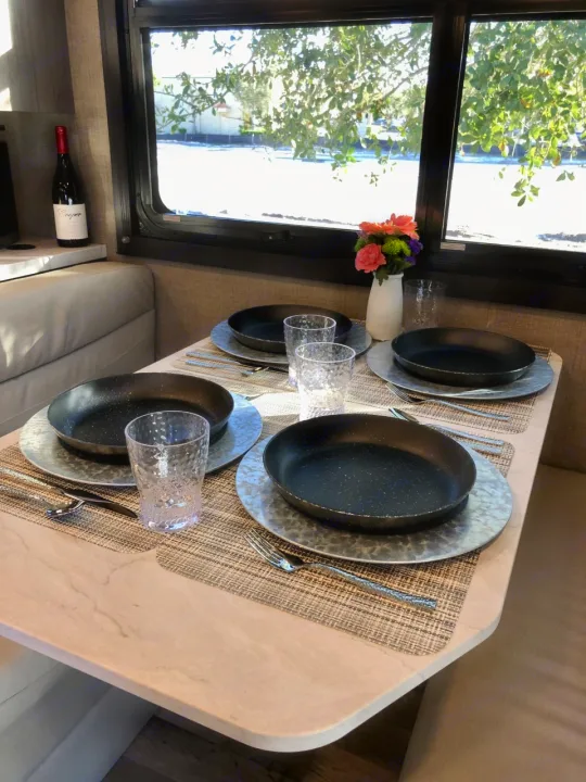 RV rental dinette: Stylish table set with dark dishes, metallic chargers, clear glasses, flowers, and wine. Bright window views.. Jayco Redhawk 2023