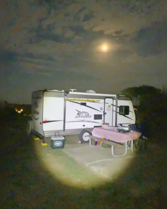 An RV rental parked at a campsite at night, illuminated by a bright moon. A picnic table and cooler are beside it.. Jayco Jay Flight SLX 2018
