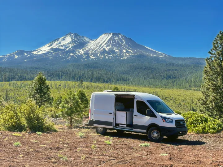 A white Ford Transit RV rental van, door open, sits on dirt, facing snow-capped Mount Shasta & forested valley under blue sky.. Ford Transit 2020