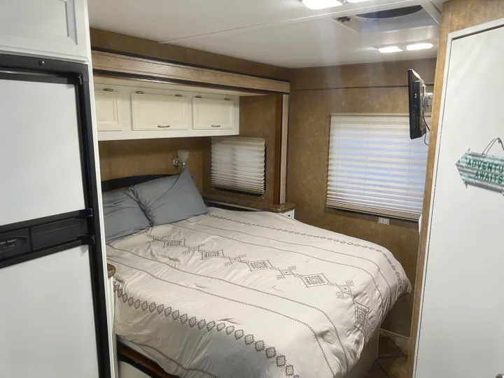 Cozy RV rental bedroom featuring a patterned bed, overhead cabinets, wall-mounted TV, and blinds.. Thor Four Winds 2014