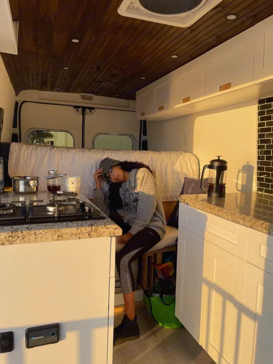 Inside a cozy RV rental: a woman relaxes in the sun-drenched kitchen area with stove, French press, and wooden ceiling.. Other Other 2020