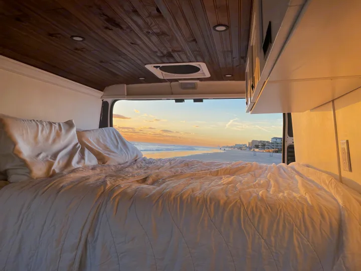Cozy RV rental bed with pillows, offering stunning ocean views of a tranquil beach, waves, and a vibrant sunset sky.. Other Other 2020