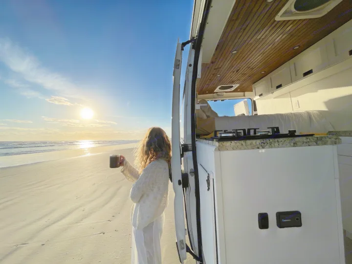 Woman sips coffee, watching a beach sunrise from her RV rental. Campervan open to sandy shore.. Other Other 2020