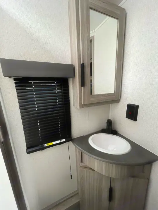 Inside an RV rental: a compact bathroom with a corner sink, mirrored medicine cabinet, and black window blinds.. Keystone Passport 2022