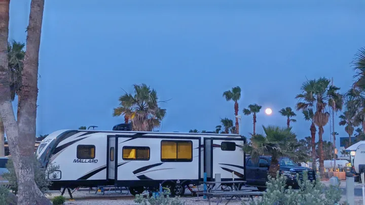 Night view of a parked Mallard travel trailer RV rental at a palm-lined campground under a full moon, lights on inside.. Heartland RVs Mallard 2019