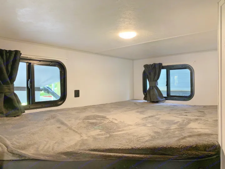 Cozy RV rental sleeping area featuring a wide bed with a soft grey blanket, two windows with dark curtains, and an overhead light.. Forest River Salem FSX 2021