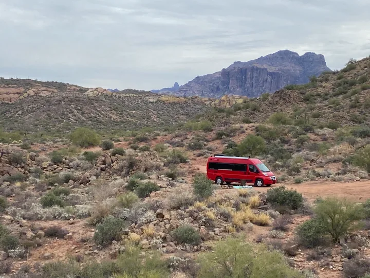 A red RV rental is parked for camping in a vast desert landscape with distant rugged mountains and sparse vegetation.. Winnebago Travato 2015