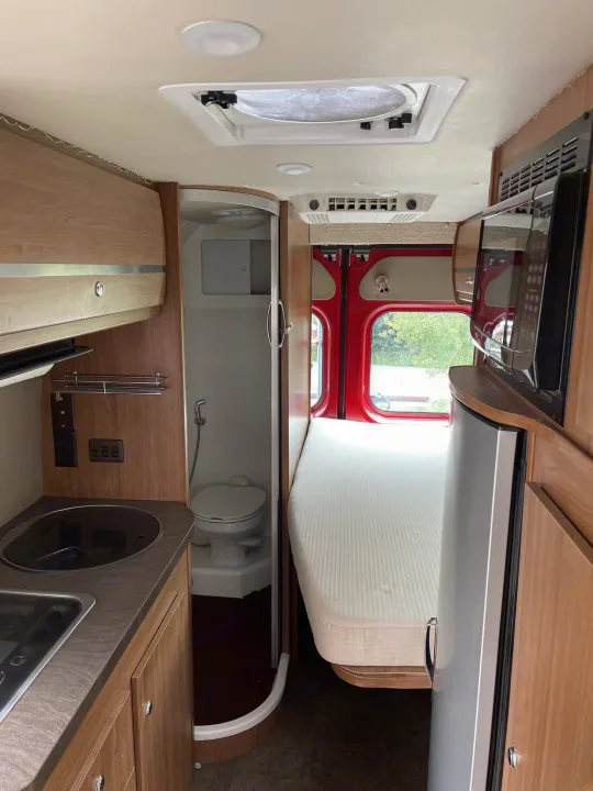 Explore this RV rental's compact interior: kitchen with sink/cooktop, microwave, fridge, toilet/shower, and bed area.. Winnebago Travato 2015