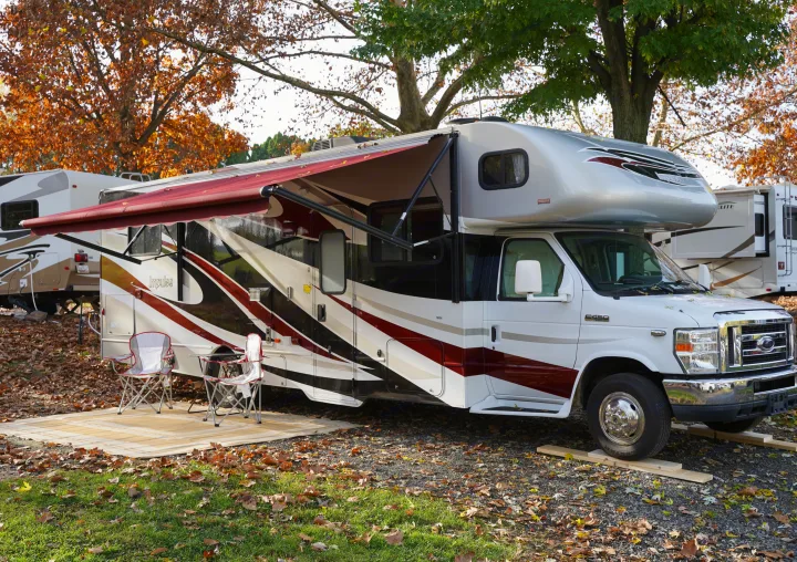 White and maroon Class C rv rental parked at an autumn campsite. Awning extended, chairs on a rug amidst fallen leaves.. Itasca Impulse 2013
